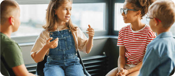 A group of children are gathered, with a girl in overalls animatedly speaking to her peers in a well-lit room.